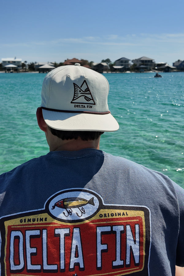 Person wearing a 'Delta Fin Bait & Tackle' shirt and cap by a body of water.