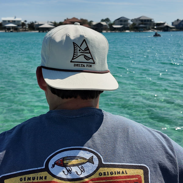 Person wearing a 'Delta Fin Bait & Tackle' shirt and cap by a body of water.
