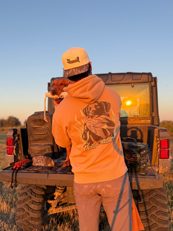 Person standing next to a truck in a field with a dog on their shoulder, wearing an orange hoodie with a bear design.