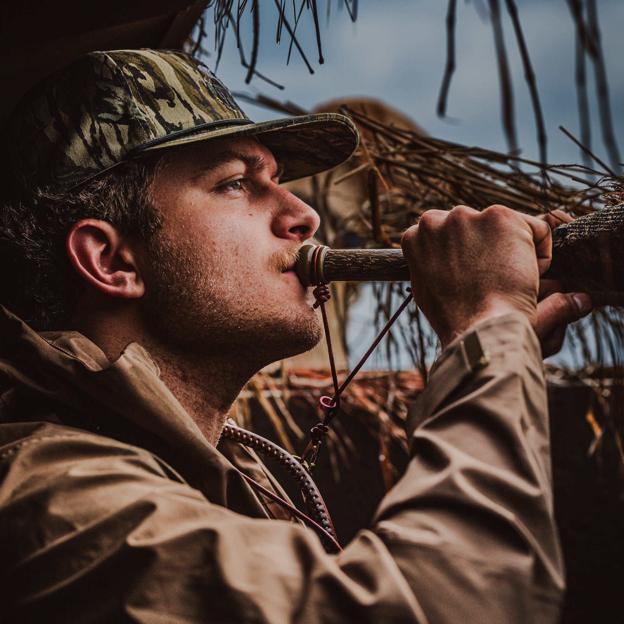 Man in camouflage hat and jacket blowing a hunting call in a natural setting