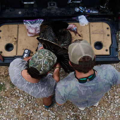 Two people in camouflage clothing standing next to a large turkey in the back of a truck.