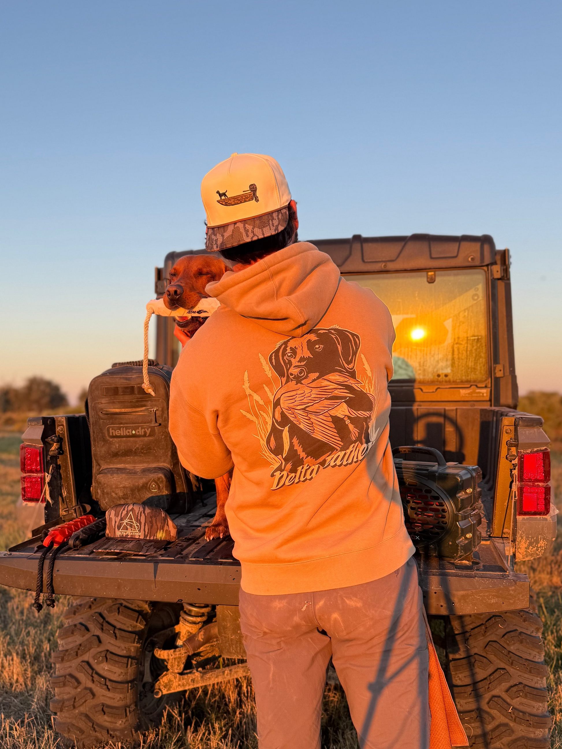 Person standing next to a truck in a field with a dog on their shoulder, wearing an orange hoodie with a bear design.