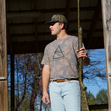 Man holding a rifle, wearing camouflage clothing and a cap, standing inside a wooden structure with trees outside.